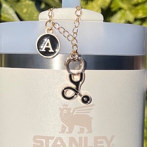 Puede incluir: Una cadena dorada con un dije de letra A negro y dorado y un dije de estetoscopio negro y dorado unidos a una taza de viaje Stanley blanca.