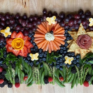 May include: A colorful charcuterie board with a garden theme. The board features a variety of fruits, vegetables, and cheeses arranged in the shape of flowers. There are red grapes, blueberries, strawberries, carrots, and cheese. The board is decorated with edible butterflies and ladybugs.