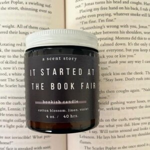 May include: A dark glass jar candle with a white lid, featuring the text "IT STARTED AT THE BOOK FAIR" in white. The label also includes "bookish candle" and scent details: cotton blossom, linen, and violet. The candle is 4 oz. and burns for 40 hours.