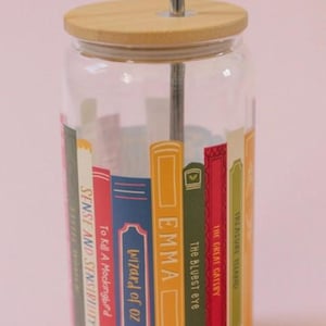 May include: A clear glass tumbler with a bamboo lid and a silver straw. The tumbler is decorated with colorful book spines featuring titles like "Emma" and "The Great Gatsby." A fun and literary-themed drinkware item.