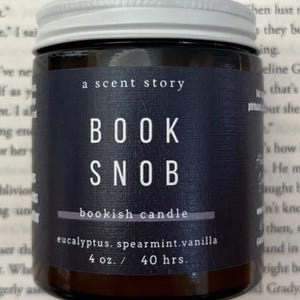 May include: A dark glass jar with a silver lid, labeled "BOOK SNOB" with "bookish candle" below. The label also lists eucalyptus, spearmint, and vanilla scents. The candle is 4 oz and has a 40-hour burn time.