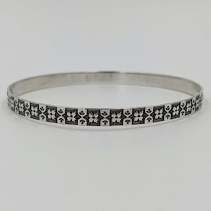 May include: A silver-colored bangle bracelet with a repeating pattern of dark, stylized floral designs. The bracelet is circular and appears to be made of metal. The design is etched into the surface, creating a contrast between the silver and the darker pattern.