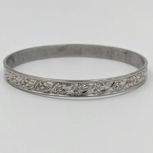 Puede incluir: Brazalete plateado con un patrón floral grabado. El brazalete tiene un diseño circular con un patrón detallado y repetitivo alrededor de la banda. La joya se muestra sobre un fondo blanco.