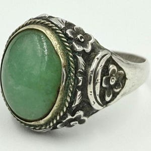 Könnte beinhalten: Ein Silberring mit einem grünen Edelstein, der in einem floralen Design gefasst ist. Der Ring hat eine breite Band mit aufwendigen Details.