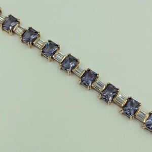 May include: A gold-toned bracelet featuring alternating square and rectangular blue-purple gemstones. The square stones are set between rectangular, clear stones, creating a repeating pattern. The bracelet is displayed against a light background.
