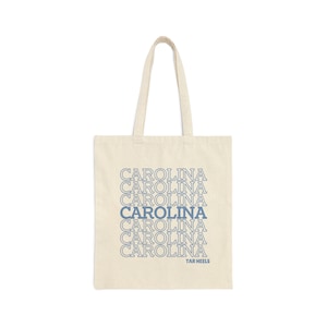 Könnte beinhalten: Eine beige Canvas-Tragetasche mit dem Wort "CAROLINA" in einem gestapelten Muster in blauer Schrift wiederholt. Der Text "TAR HEELS" ist in kleinerer blauer Schrift am unteren Rand der Tasche aufgedruckt.