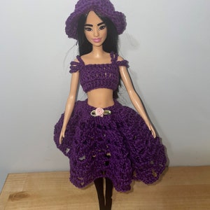 May include: A purple crocheted doll outfit with a matching hat. The outfit includes a crop top and a skirt with a pink flower accent.