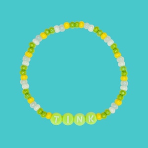 Op de afbeelding: Een kralenarmband met het woord "TINK" in limoengroene letterkralen. De armband heeft gele, witte en groene kralen, tegen een heldere turkooizen achtergrond. Een kleurrijk accessoire.