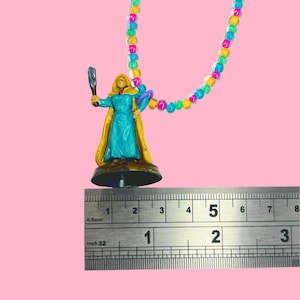 May include: A miniature figurine of a woman in a teal dress and gold cape, holding a staff and a purple object. The figurine is attached to a colorful beaded necklace. The ruler shows the figurine is approximately 2 inches tall.