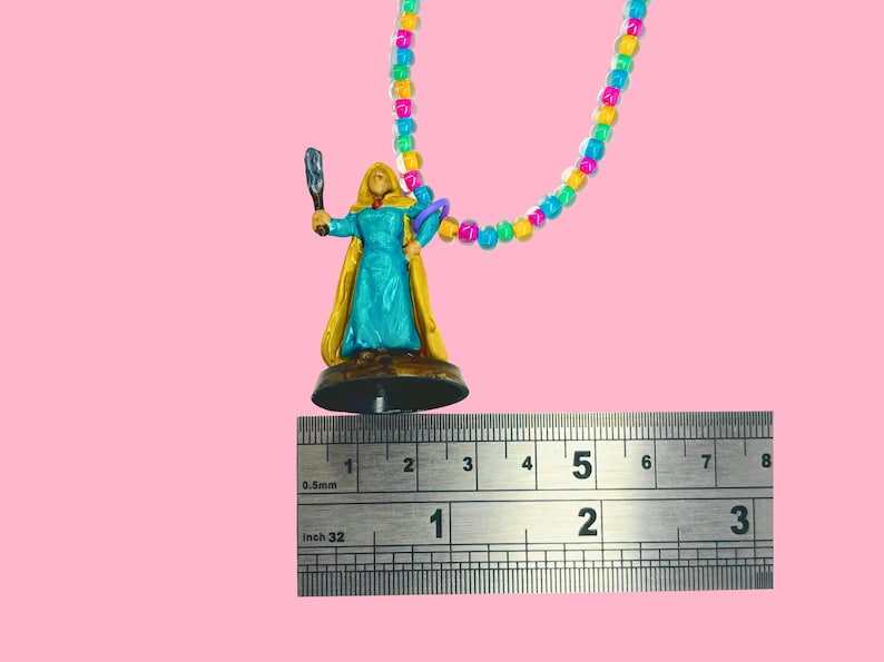 May include: A miniature figurine of a woman in a teal dress and gold cape, holding a staff and a purple object. The figurine is attached to a colorful beaded necklace. The ruler shows the figurine is approximately 2 inches tall.