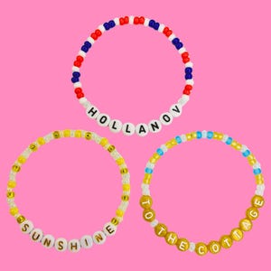 Könnte beinhalten: Drei Armbänder mit Perlen mit weißen Buchstabenperlen mit den Wörtern "HOLLANOV", "SUNSHINE" und "TO THE COTTAGE". Die Armbänder haben rote, weiße, blaue, gelbe und goldene Perlen. Der Hintergrund ist einfarbig rosa.