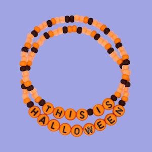 May include: Two beaded bracelets in orange, black, and cream colors. The bottom bracelet spells out "HALLOWEEN" in orange letter beads. The top bracelet has the words "THIS IS" in orange letter beads. The bracelets are on a light purple background.
