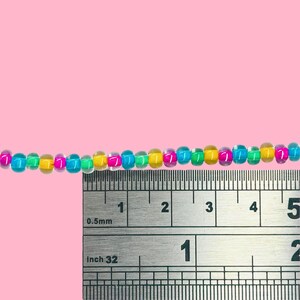 May include: A strand of colorful beads, approximately 5 inches long, in shades of blue, green, yellow, pink, and purple. The beads are cylindrical and are next to a ruler for scale. The background is a solid pink.