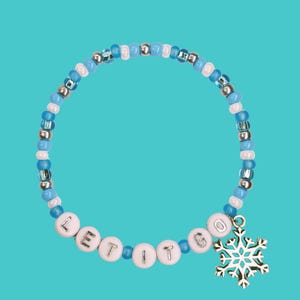 Könnte beinhalten: Ein Armband aus blauen, weißen und silbernen Perlen, mit den Buchstaben "LET IT GO" aus weißen Buchstabenperlen. Es hat einen silbernen Schneeflocken-Anhänger. Das Armband wird auf einem türkisfarbenen Hintergrund präsentiert.