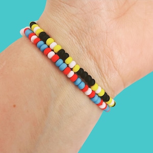 May include: Three beaded bracelets in red, white, blue, yellow, and black are stacked on a wrist. The bracelets are made of small, round beads. The background is a solid teal color.