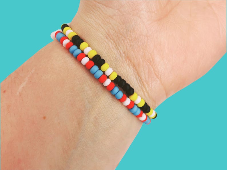 May include: Three beaded bracelets in red, white, blue, yellow, and black are stacked on a wrist. The bracelets are made of small, round beads. The background is a solid teal color.
