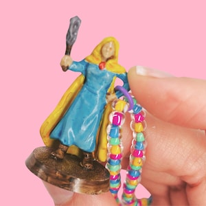 May include: A miniature figurine of a robed figure in blue and yellow, holding a torch. The figurine is on a brown base. A colorful beaded bracelet is held near the figurine. The background is pink.
