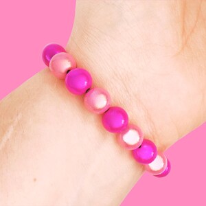 A close-up of a wrist wearing a pink beaded bracelet with shiny, round beads, set against a solid pink background.