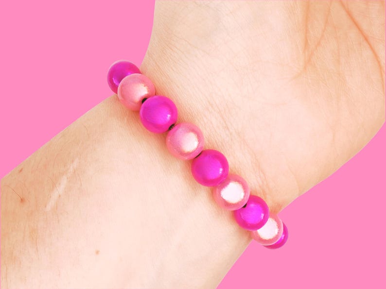 A close-up of a wrist wearing a pink beaded bracelet with shiny, round beads, set against a solid pink background.