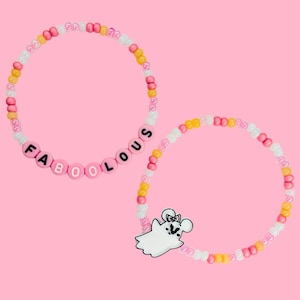 Może przedstawiać: Dwie bransoletki z koralików na różowym tle. Jedna bransoletka wymawia "FABOOLOUS" w różowych koralikach z literami. Druga bransoletka ma zawieszkę z duchem z kokardą. Bransoletki wykonane są z różowych, białych i żółtych koralików.