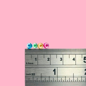 May include: Four small, colorful beads in blue, green, yellow, and pink are displayed on a ruler. The ruler shows measurements in inches and millimeters. The beads are approximately 0.125 inches in diameter.