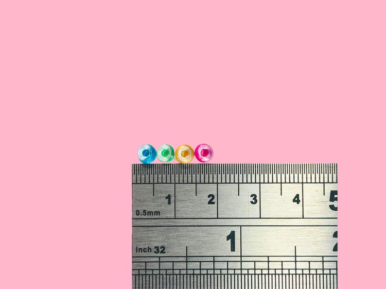 May include: Four small, colorful beads in blue, green, yellow, and pink are displayed on a ruler. The ruler shows measurements in inches and millimeters. The beads are approximately 0.125 inches in diameter.