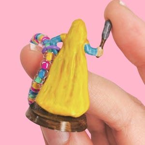 May include: A miniature figurine of a person in a yellow gown, holding a small weapon. The figure has blue sleeves and is on a brown base. A colorful beaded bracelet is also visible. The background is a solid pink.