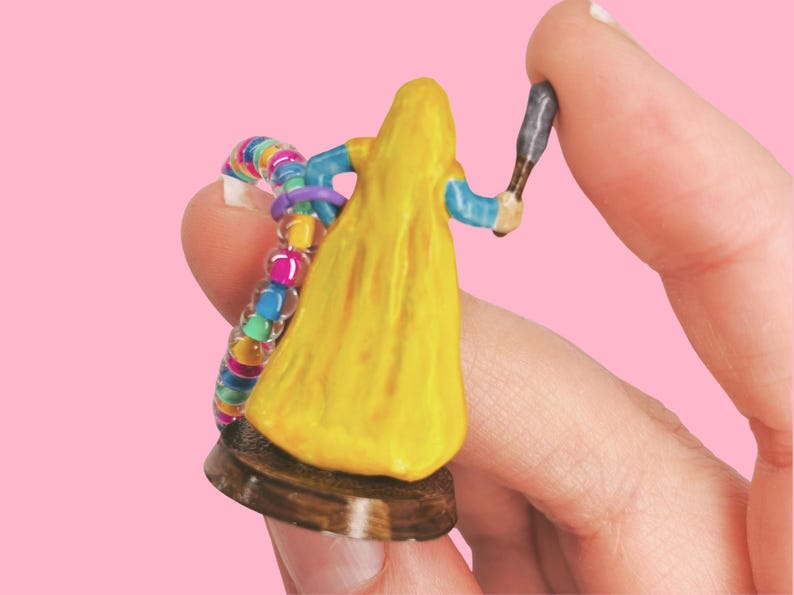 May include: A miniature figurine of a person in a yellow gown, holding a small weapon. The figure has blue sleeves and is on a brown base. A colorful beaded bracelet is also visible. The background is a solid pink.