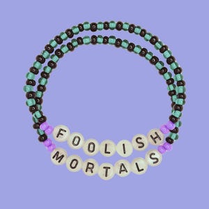 May include: A black and teal beaded bracelet with white beads that spell out "FOOLISH MORTALS".