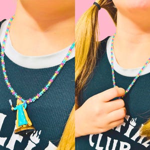 May include: A colorful beaded necklace with a pendant featuring a figure in a blue dress and gold cape. The necklace is made of small, multi-colored beads. The figure is standing on a brown base. The necklace is worn over a dark blue tank top.