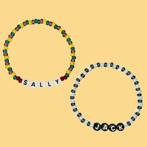 Puede incluir: Dos pulseras de cuentas con nombres escritos en letras blancas. La primera pulsera es roja, amarilla y verde con el nombre "SALLY". La segunda pulsera es negra y blanca con el nombre "JACK".