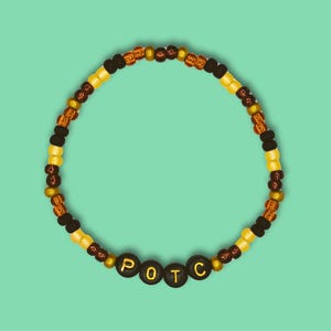 Puede incluir: Un brazalete de cuentas marrones, doradas y negras con las letras "POTC" escritas en cuentas negras.