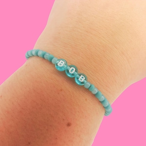 May include: A turquoise beaded bracelet spelling out "BOB" with white letter beads. The bracelet is worn on a wrist against a pink background. The beads are small and round.