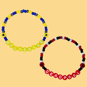 Pode incluir: Duas pulseiras de contas, uma amarela com a palavra "WOLVERINE" e a outra vermelha com a palavra "DEADPOOL".