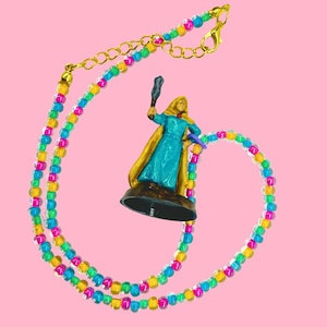May include: A colorful beaded necklace with a gold clasp and a miniature figurine. The necklace features a pattern of yellow, pink, blue, and green beads. The figurine is of a person in a blue dress and gold cape, holding a staff.