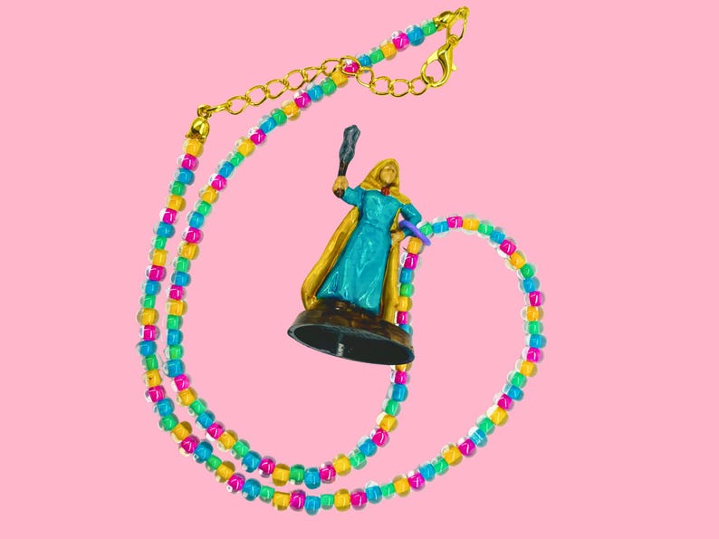 May include: A colorful beaded necklace with a gold clasp and a miniature figurine. The necklace features a pattern of yellow, pink, blue, and green beads. The figurine is of a person in a blue dress and gold cape, holding a staff.