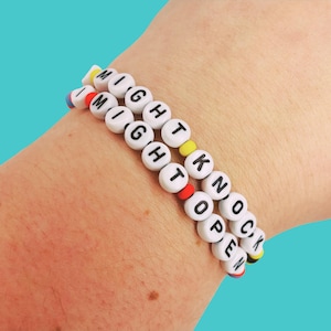 May include: Two beaded bracelets spelling "I MIGHT KNOCK OPEN" in white letter beads with colorful accents. The bracelets are worn on a wrist against a teal background.