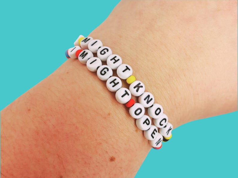 May include: Two beaded bracelets spelling "I MIGHT KNOCK OPEN" in white letter beads with colorful accents. The bracelets are worn on a wrist against a teal background.
