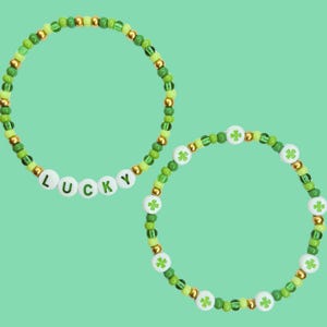 May include: Two beaded bracelets in shades of green and gold. One bracelet spells "LUCKY" with white letter beads. The other bracelet features white beads with green shamrock designs. The bracelets are on a light green background.