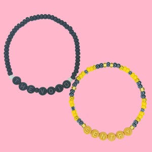 May include: Two beaded bracelets on a pink background. One bracelet is black with the text "THE VOID" in white letters. The other bracelet is yellow and black with the text "SENTRY" in gold letters.