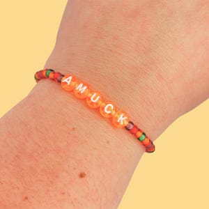 May include: A beaded bracelet spelling "AMUCK" in white letters on orange beads. The bracelet has a rainbow-colored band. The bracelet is worn on a person's wrist.