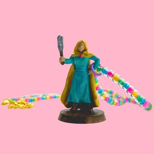 May include: A miniature figurine of a robed figure in teal and gold, holding a weapon and a colorful beaded chain. The figure stands on a brown base against a pink background. A second beaded chain lies on the left.