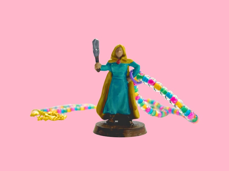 May include: A miniature figurine of a robed figure in teal and gold, holding a weapon and a colorful beaded chain. The figure stands on a brown base against a pink background. A second beaded chain lies on the left.