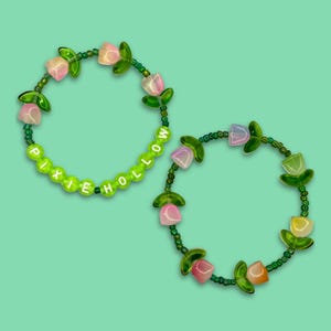 May include: Two beaded bracelets with green and pink flower charms. One bracelet spells out "PIXIE HOLLOW" in white letters.