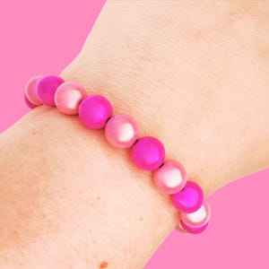 A wrist wearing a bracelet made of shiny, round pink beads, set against a solid pink background.