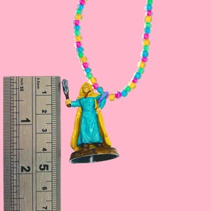 May include: A colorful beaded necklace with a miniature figurine. The figurine is a woman in a blue dress and yellow cape, holding a weapon. The necklace is approximately 1 inch in length.