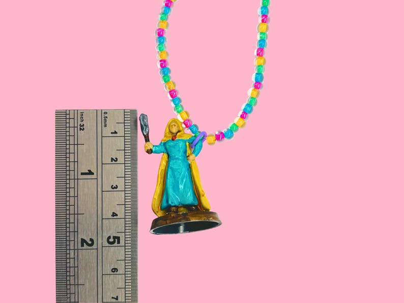 May include: A colorful beaded necklace with a miniature figurine. The figurine is a woman in a blue dress and yellow cape, holding a weapon. The necklace is approximately 1 inch in length.
