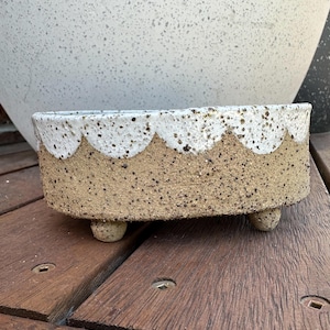 Peut inclure: Un pot en céramique blanc et brun avec un bord festonné et trois petits pieds. Le pot a un glaçage moucheté.