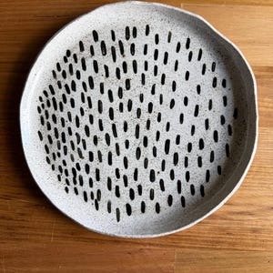 May include: A round ceramic plate with an off-white speckled finish and a slightly irregular, hand-formed edge. The surface is decorated with a repeating pattern of short, vertical black dashes arranged in a dense, scattered formation across the centre.