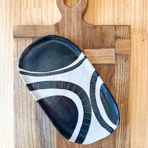 Op de afbeelding: Een handgemaakte, ovale keramische serveerschaal met een zwart-wit abstract ontwerp. De schaal ligt op een houten snijplank met een handvat. De schaal is ongeveer 30 cm lang en 15 cm breed.